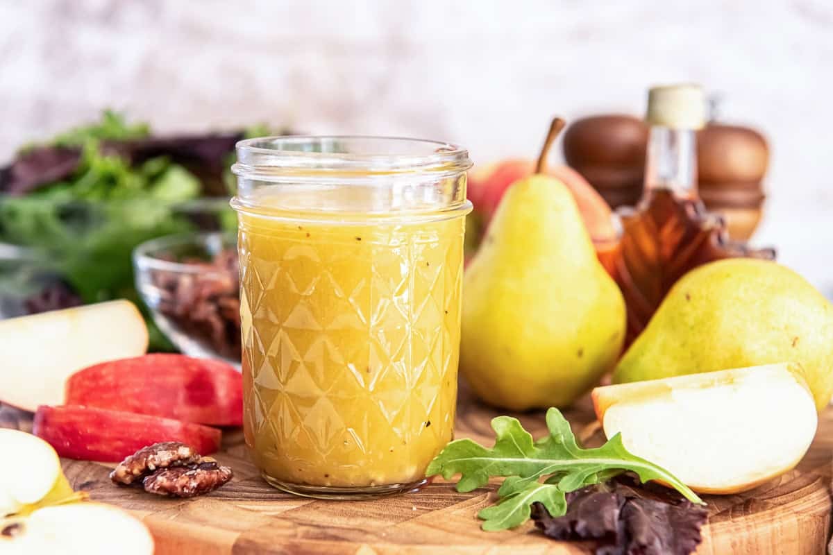 A jar of Maple Dijon Vinaigrette on a wooden board surrounded by apple slices, pecans, greens, pears, and a bottle of maple syrup.