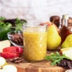 A jar of Maple Dijon Vinaigrette on a wooden board surrounded by apple slices, pecans, greens, pears, and a bottle of maple syrup.
