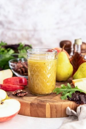A jar of Maple Dijon Vinaigrette on a wooden board surrounded by apple slices, pecans, greens, pears, and a bottle of maple syrup.