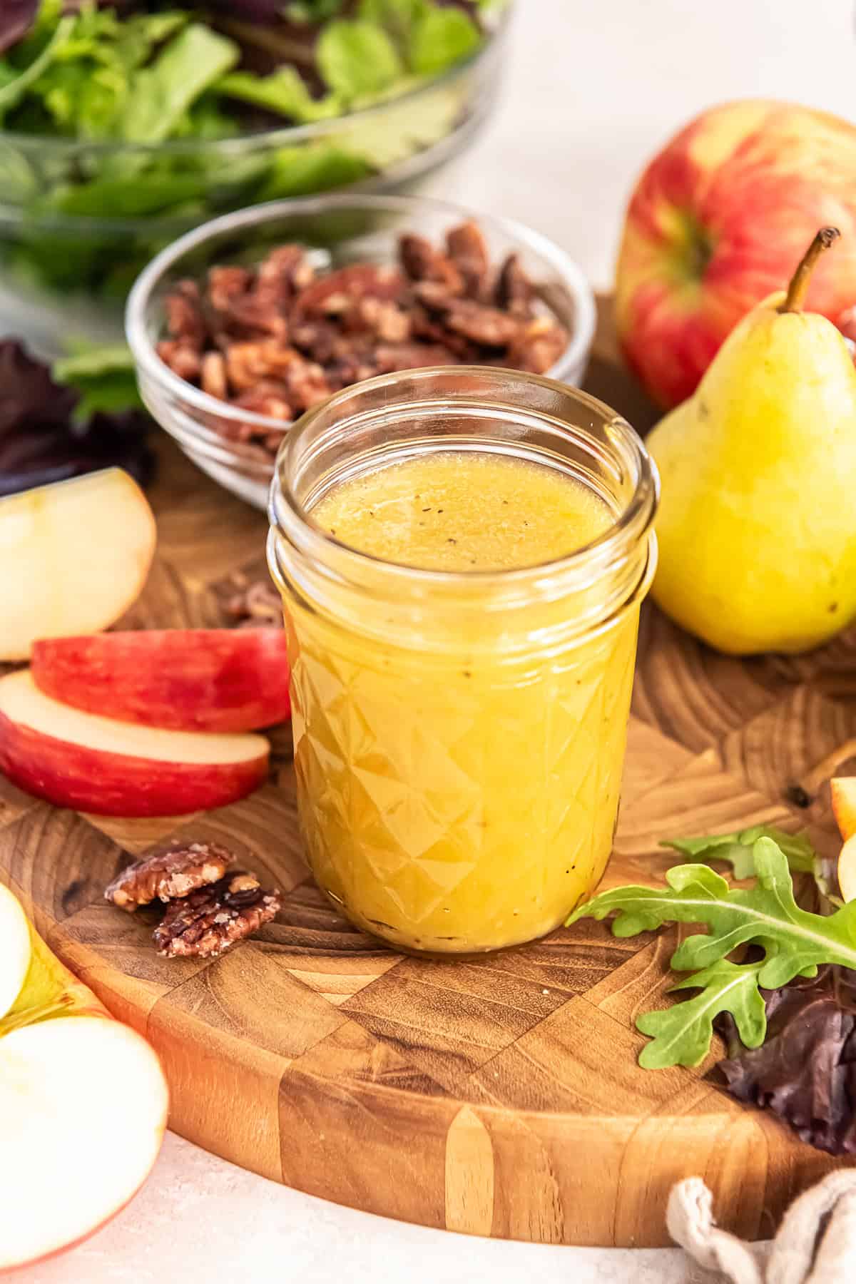 Top-down view of a jar of Maple Dijon Vinaigrette on a wooden board with candied pecans, apples, greens, and pears.