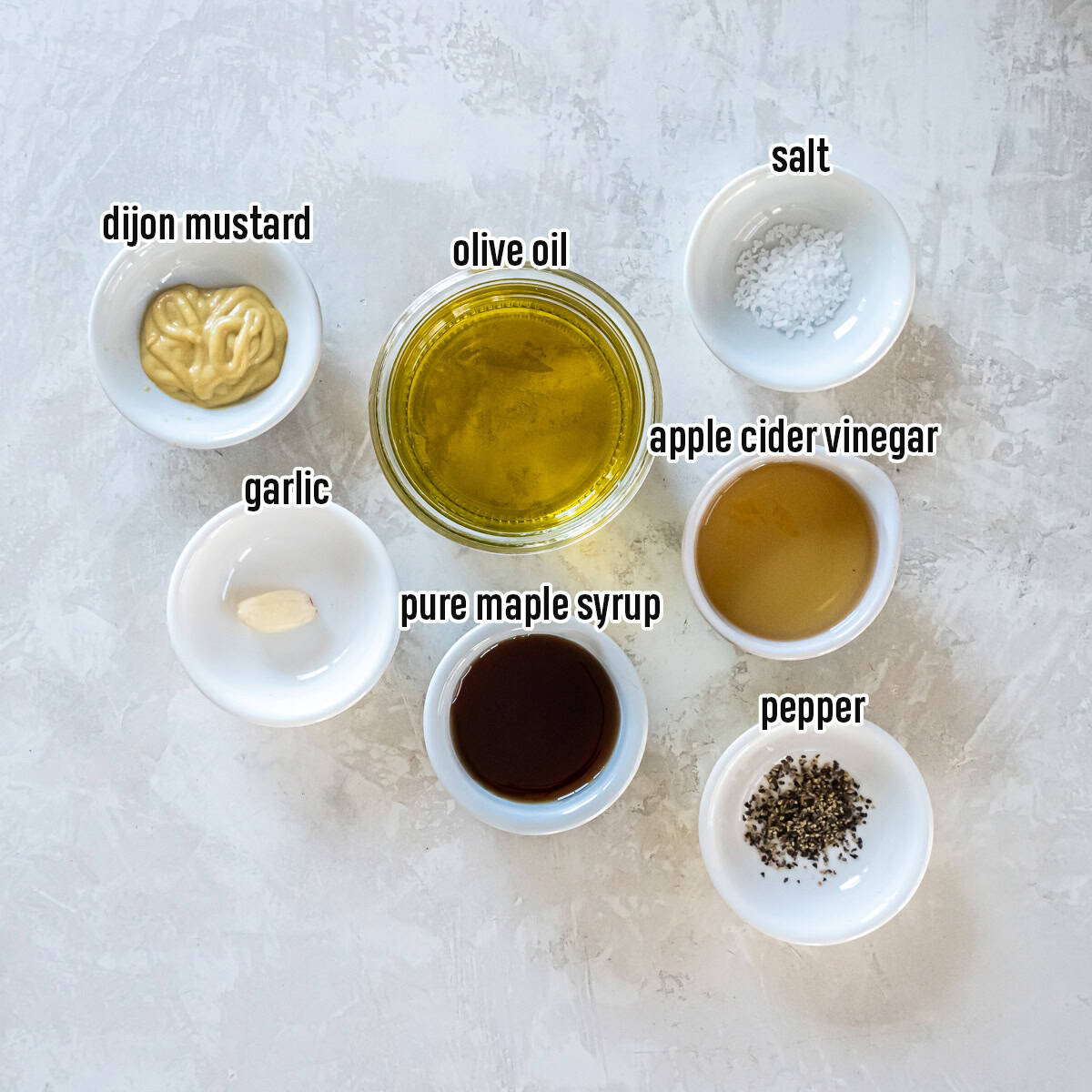 Ingredients for Maple Dijon Vinaigrette arranged in small bowls including olive oil, apple cider vinegar, maple syrup, Dijon mustard, garlic, salt, and pepper.