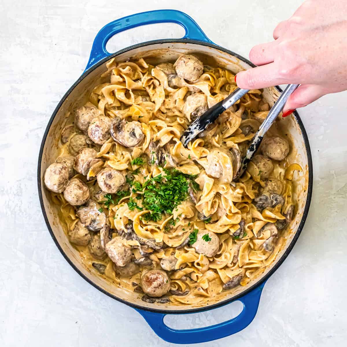 A hand with tongs mixing meatball stroganoff—egg noodles, meatballs, and creamy sauce—garnished with chopped parsley in a blue-handled skillet.