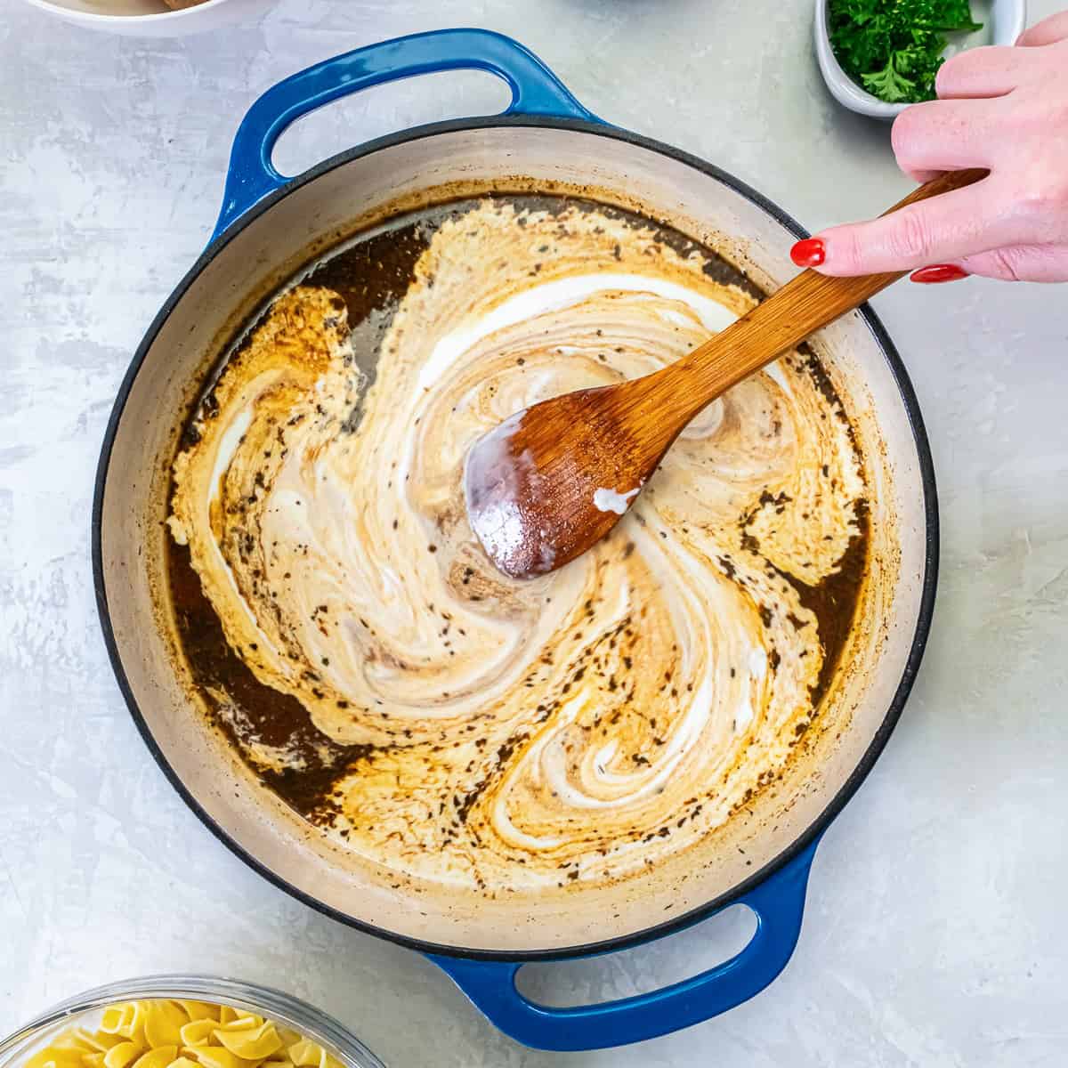 Wooden spoon swirling heavy cream into the browned pan juices to form the stroganoff sauce.