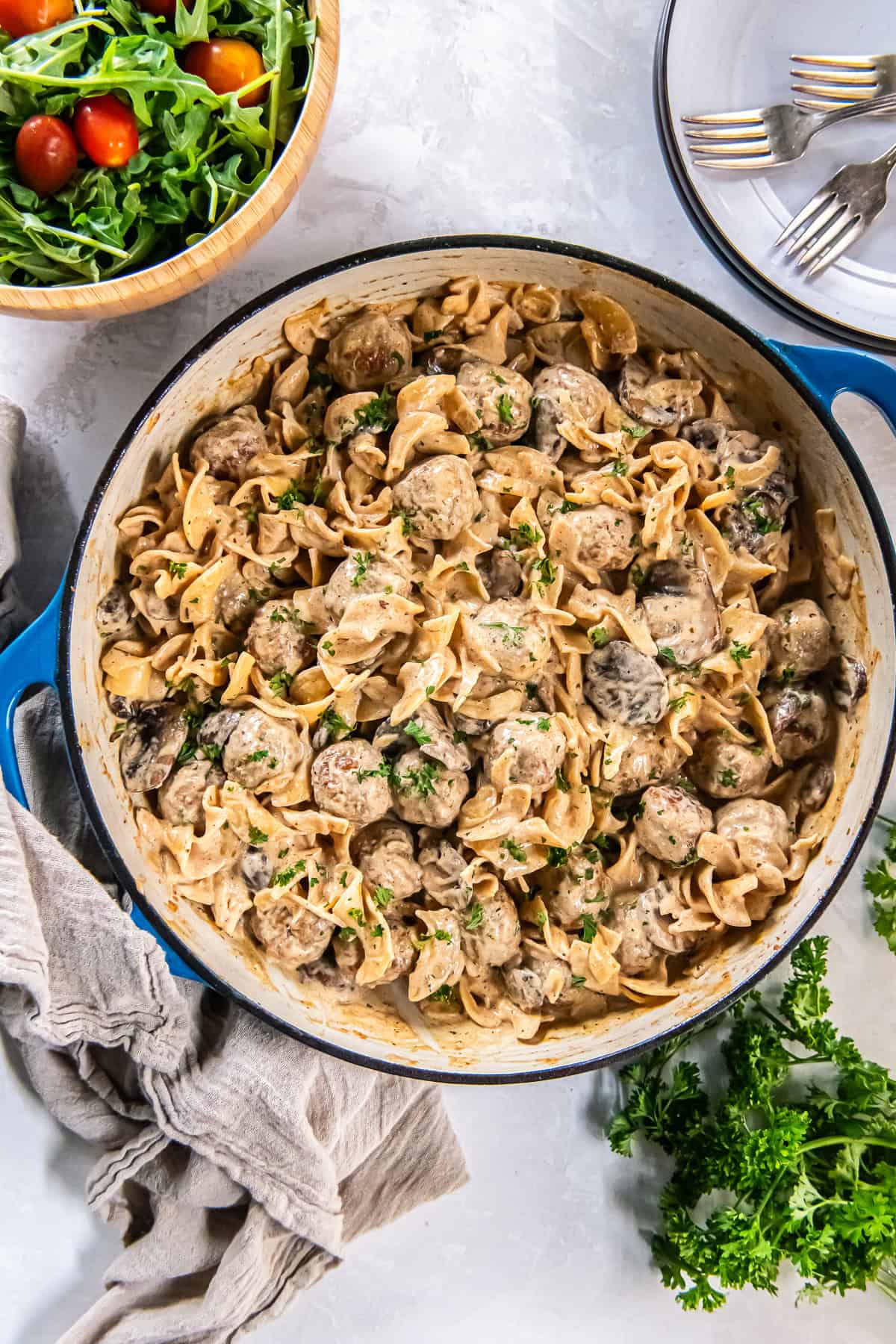 Creamy Meatball Stroganoff with wide egg noodles, mushrooms, and meatballs in a blue-handled skillet, topped with chopped parsley, with a green salad and plates with forks nearby.