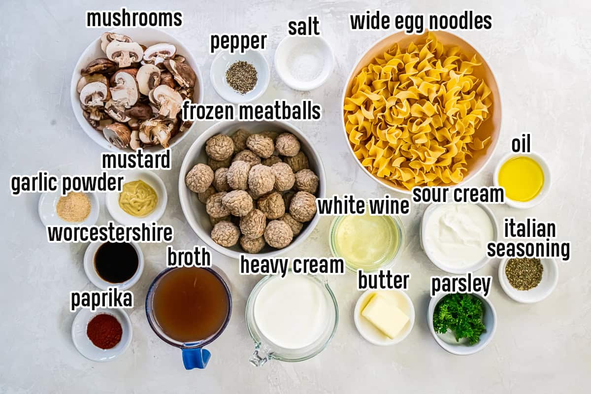 Meatball Stroganoff ingredients on a light surface: wide egg noodles, frozen meatballs, sliced cremini mushrooms, olive oil, butter, sour cream, heavy cream, white wine, beef broth, Dijon mustard, Worcestershire sauce, Italian seasoning, paprika, garlic powder, salt, pepper, and parsley.