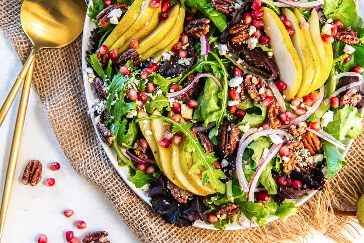 A large platter of Pear Pomegranate Salad layered with sliced pears, mixed greens, pomegranate arils, pecans, red onion, and crumbled feta over a burlap cloth.