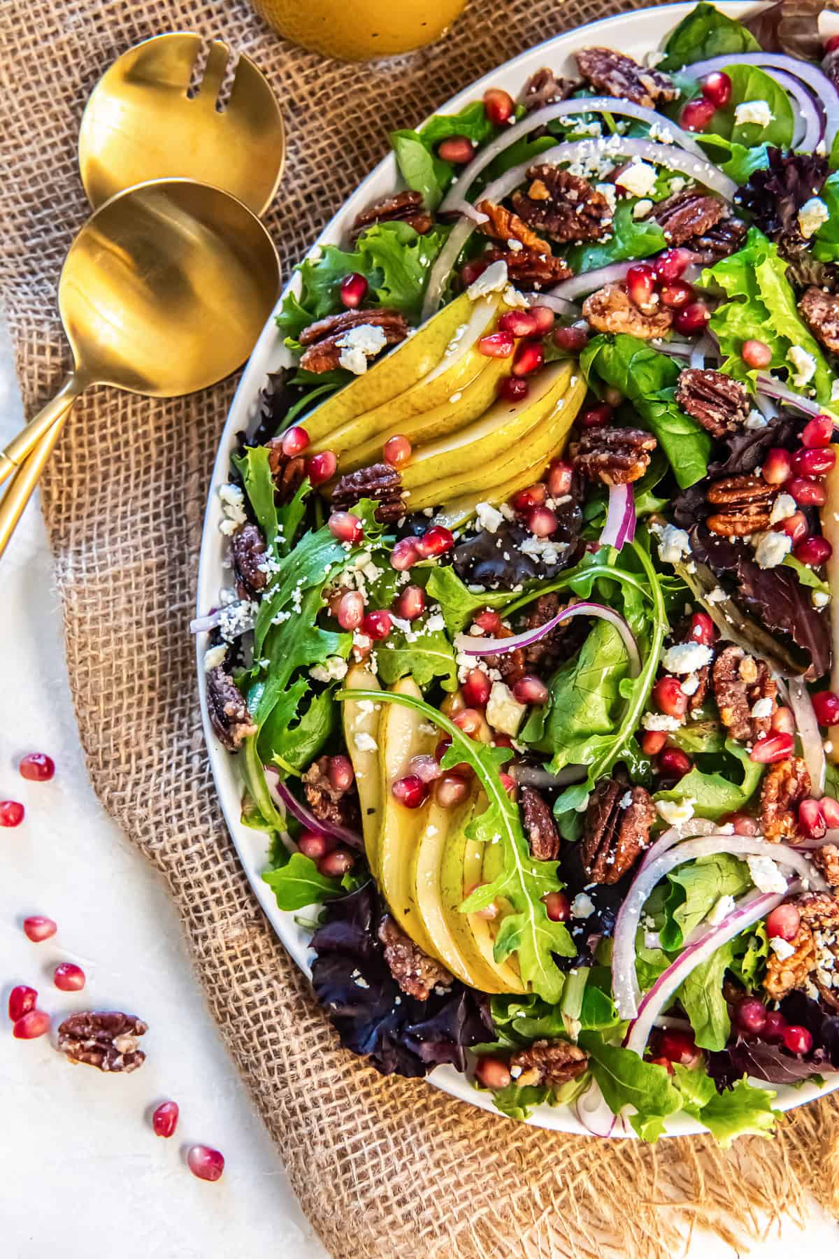 A platter of Pear Pomegranate Salad topped with sliced pears, mixed greens, pomegranate arils, pecans, red onion, and feta, styled with gold serving spoons and scattered pomegranate seeds.