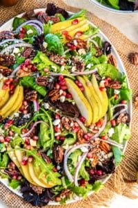A colorful Pear Pomegranate Salad with sliced pears, mixed baby greens, pomegranate arils, red onion, glazed pecans, and feta arranged on a white serving platter.