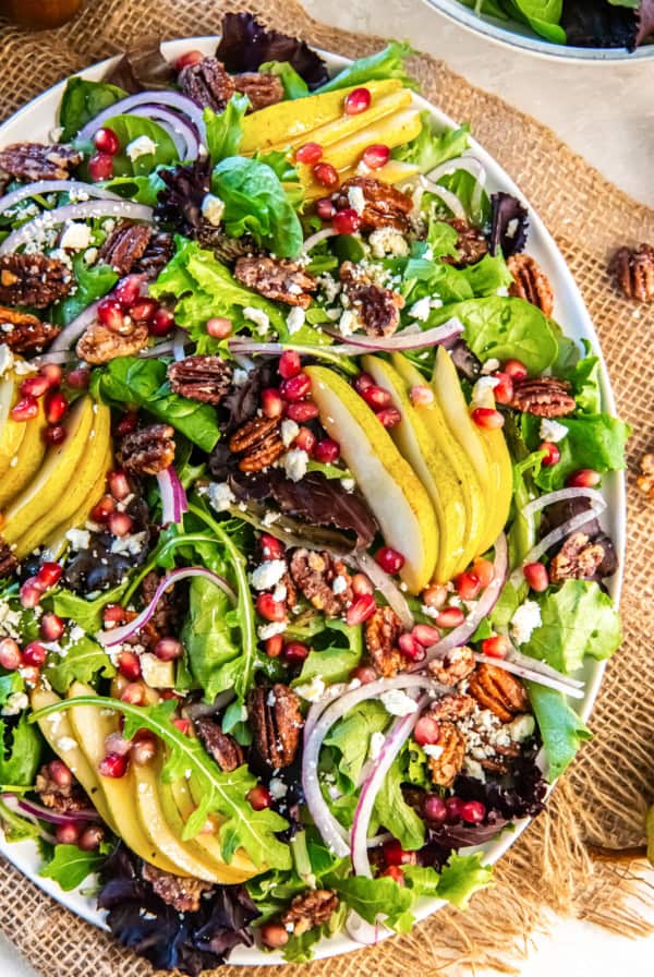 A colorful Pear Pomegranate Salad with sliced pears, mixed baby greens, pomegranate arils, red onion, glazed pecans, and feta arranged on a white serving platter.
