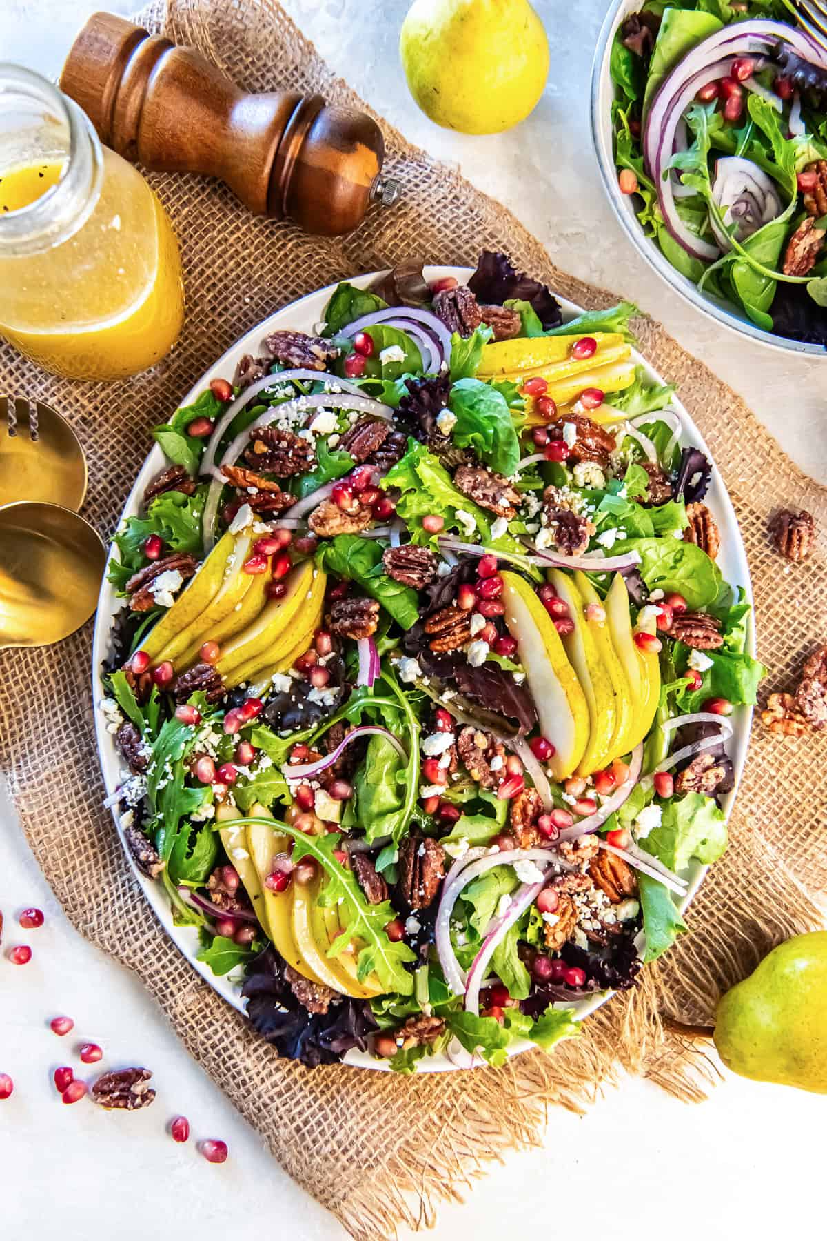 Pear Pomegranate Salad on a large platter, surrounded by a jar of dressing, salad tongs, and whole pears, featuring mixed greens, sliced pears, pomegranate arils, red onion, glazed pecans, and crumbled feta.