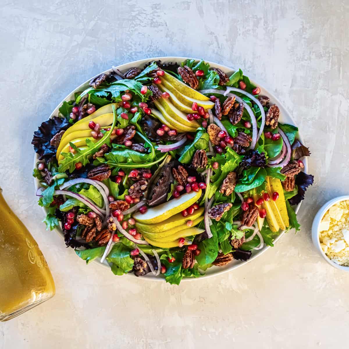 Mixed baby greens arranged on a large platter, topped with sliced pears, red onion, pomegranate arils, and glazed pecans, ready for serving.