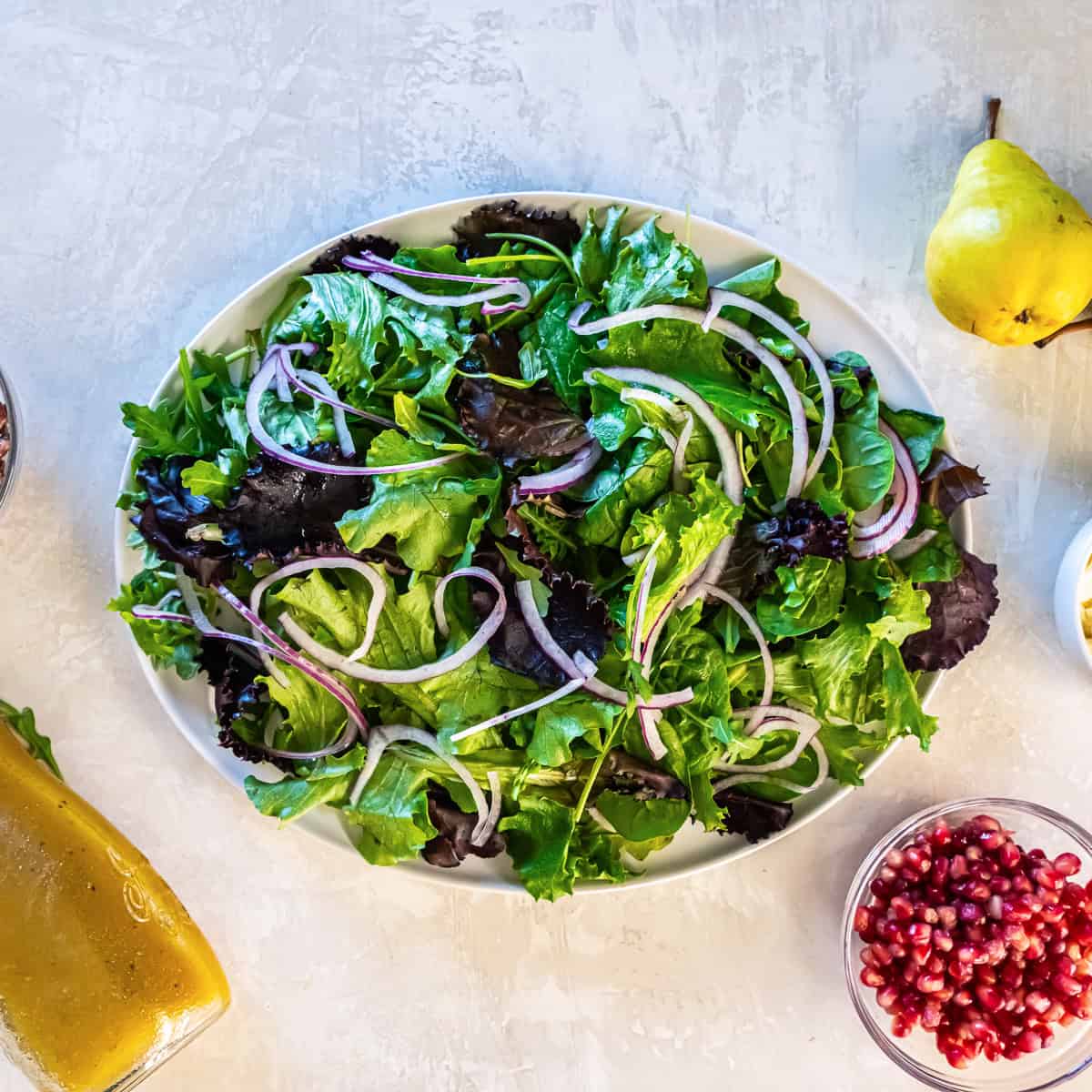 Mixed baby greens topped with thinly sliced red onion on a platter, ready for adding pears, pomegranate arils, and pecans.