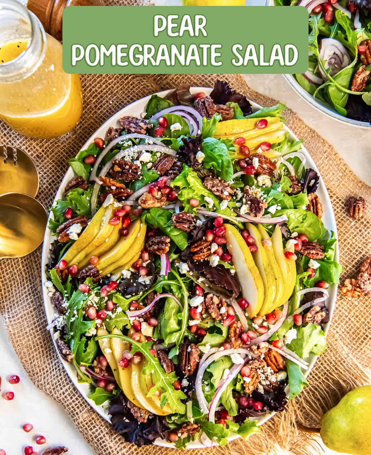 Pear Pomegranate Salad on a large platter, surrounded by a jar of dressing, salad tongs, and whole pears, featuring mixed greens, sliced pears, pomegranate arils, red onion, glazed pecans, and crumbled feta with text overlay.