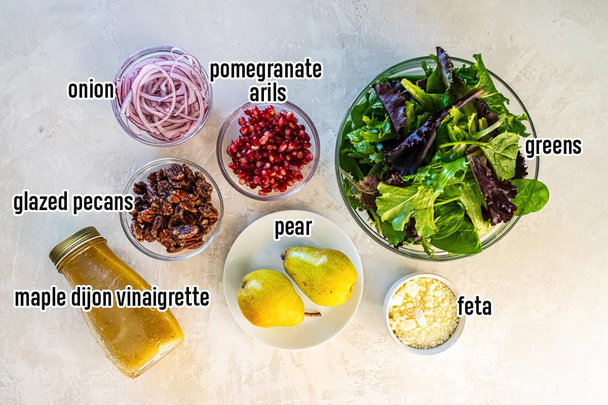Ingredients for a pear pomegranate salad arranged on a light surface, including mixed greens, sliced red onion, pomegranate arils, glazed pecans, crumbled feta, two whole pears, and a jar of maple dijon vinaigrette, each labeled with text overlay.