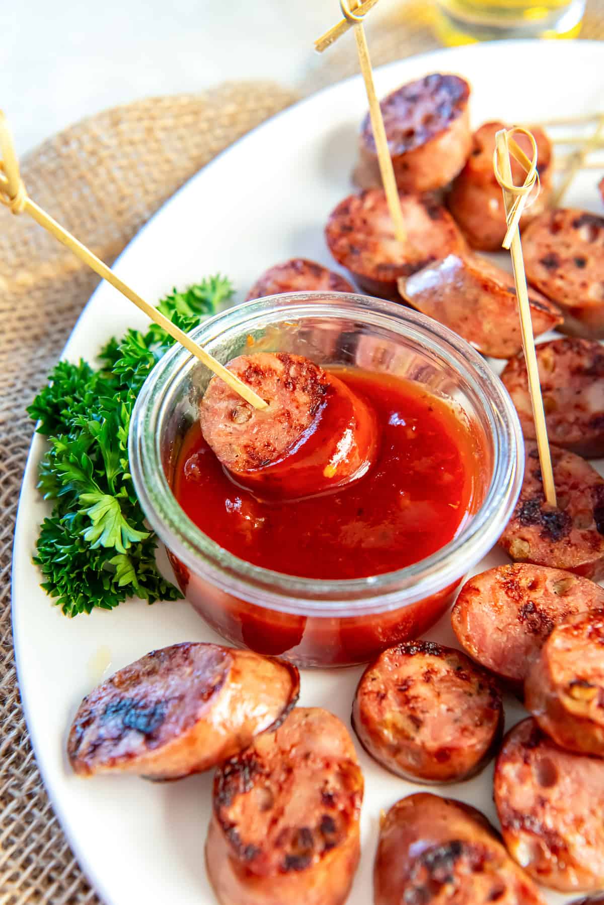Chicken sausage bite dipped into a sweet chili jam sauce in a small glass bowl, served on a white platter with toothpicks and fresh parsley.