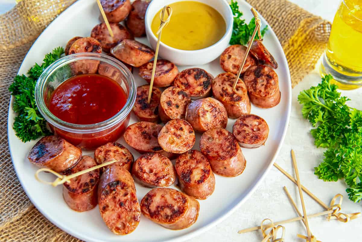 Platter of browned chicken sausage bites served with honey mustard and chili jam dipping sauces, garnished with parsley and toothpicks for easy serving.