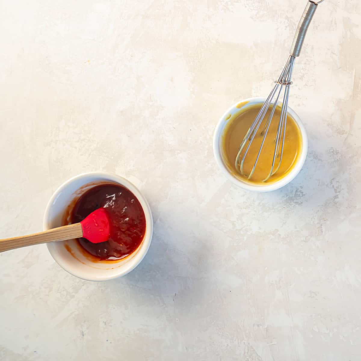 Two small bowls of dipping sauces, one with chili jam sauce and one with honey mustard being whisked on a light countertop.