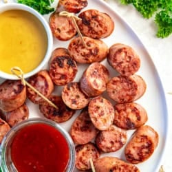 Chicken sausage bites browned in a skillet and arranged on a white platter with toothpicks, served with honey mustard and chili jam dipping sauces and fresh parsley garnish.