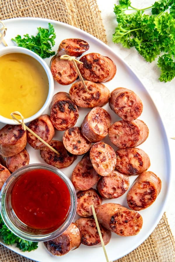 Chicken sausage bites browned in a skillet and arranged on a white platter with toothpicks, served with honey mustard and chili jam dipping sauces and fresh parsley garnish.
