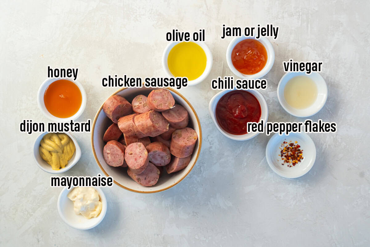 Sliced chicken sausage surrounded by small bowls of ingredients including olive oil, Dijon mustard, mayonnaise, honey, chili sauce, jam, vinegar, and red pepper flakes on a light background.