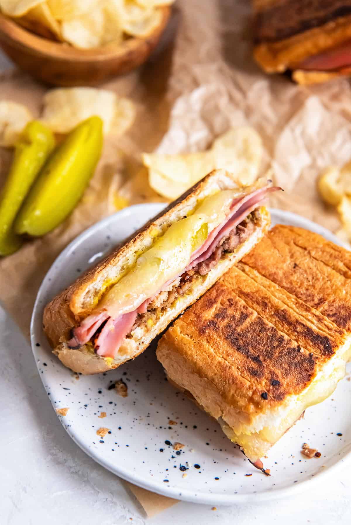 Stacked Cuban sandwich with roast pork, ham, Swiss cheese, pickles, and mustard on toasted Cuban bread, served with potato chips and dill pickles.