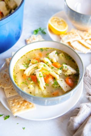 Bowl of Get Well Chicken Soup with tender chicken, carrots, and short pasta in a clear, golden broth, garnished with fresh parsley and served with saltine crackers and a lemon half in the background.