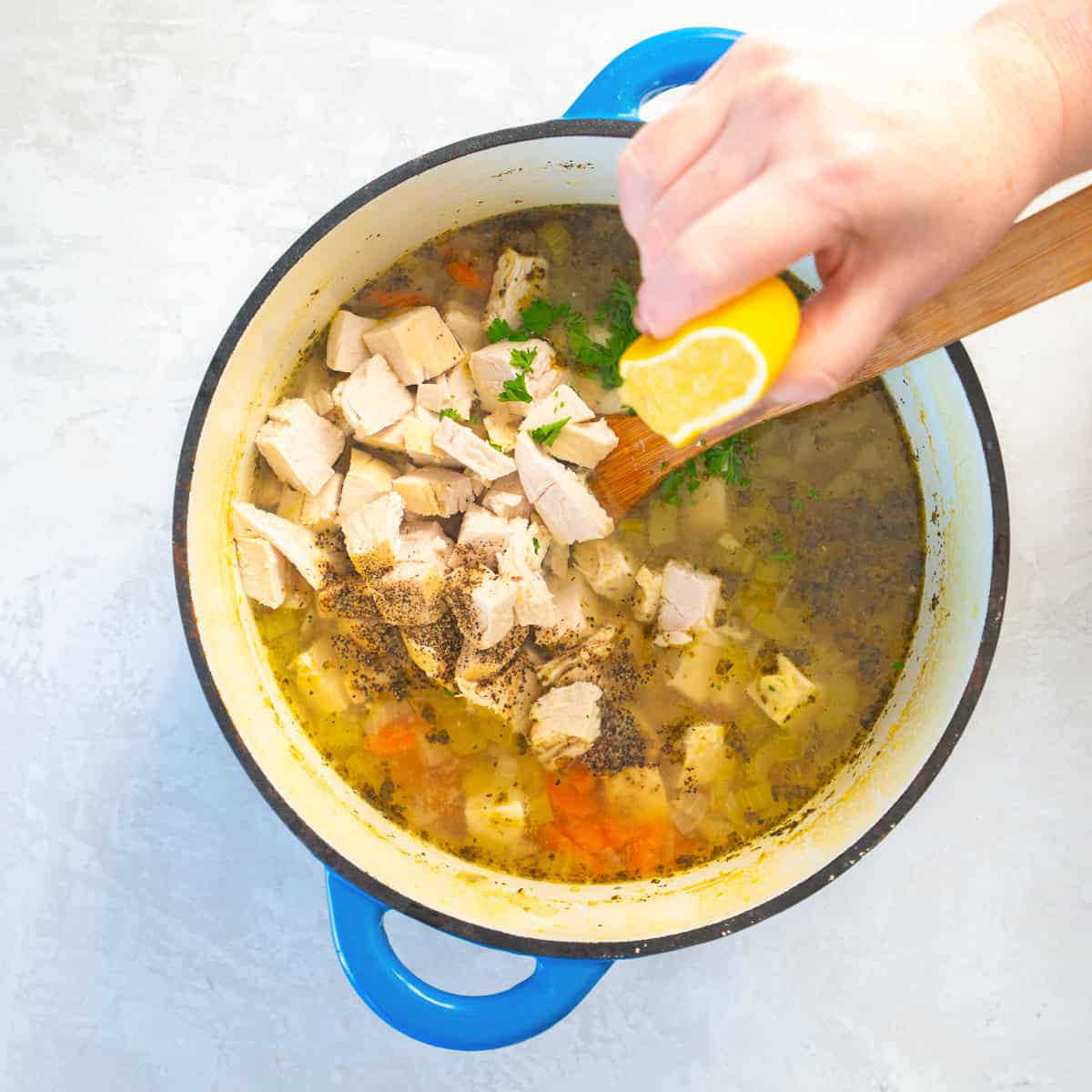 Fresh lemon juice being squeezed into a pot of chicken soup with chicken pieces and herbs.