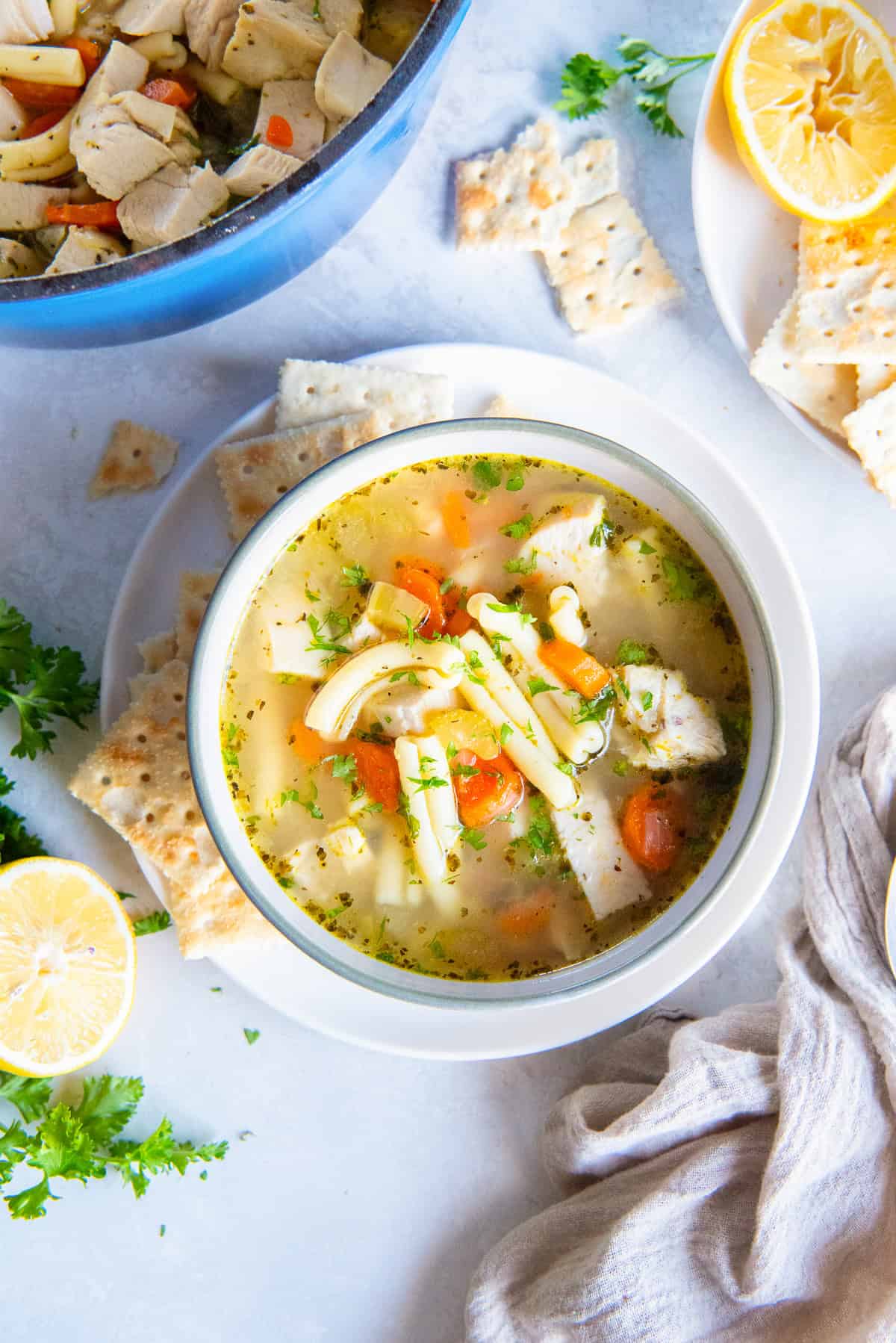 A bowl of Get Well Chicken Soup with tender chicken, carrots, short pasta, and fresh parsley in a clear broth, surrounded by saltine crackers, lemon halves, and a pot of soup in the background.