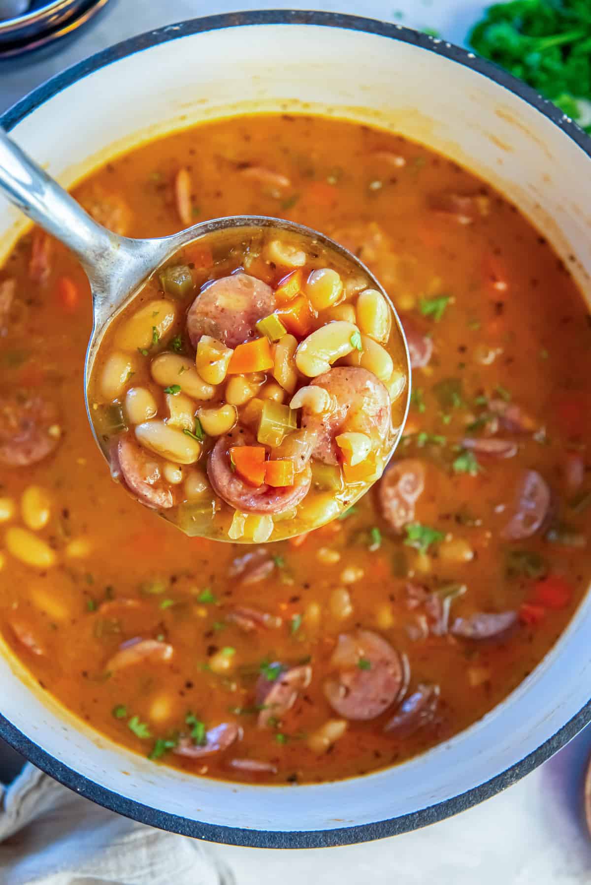 Ladle lifting sausage and bean soup from a Dutch oven, showing white beans, sliced sausage, carrots, and celery in a hearty, savory broth.