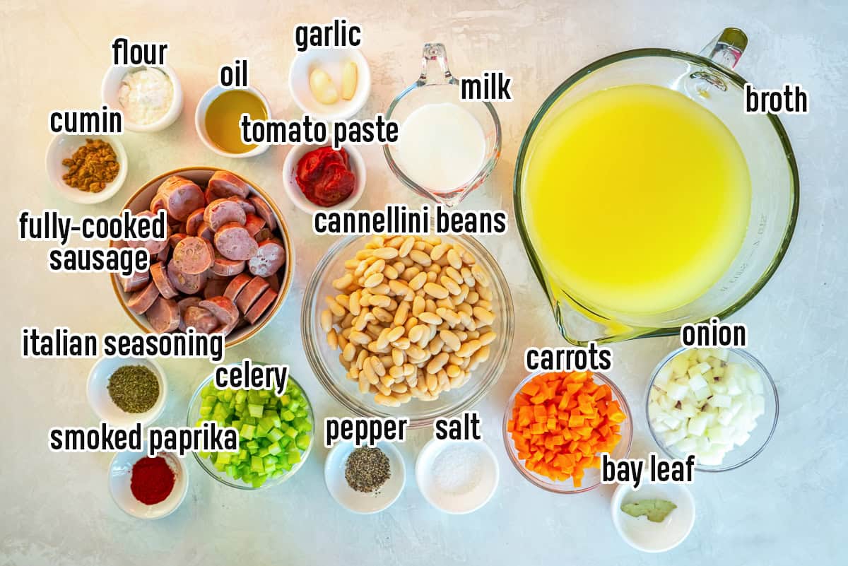 Ingredients for sausage and bean soup, including sliced chicken sausage, cannellini beans, chicken broth, onion, carrots, celery, garlic, tomato paste, milk, olive oil, flour, Italian seasoning, cumin, paprika, bay leaf, salt, and pepper.