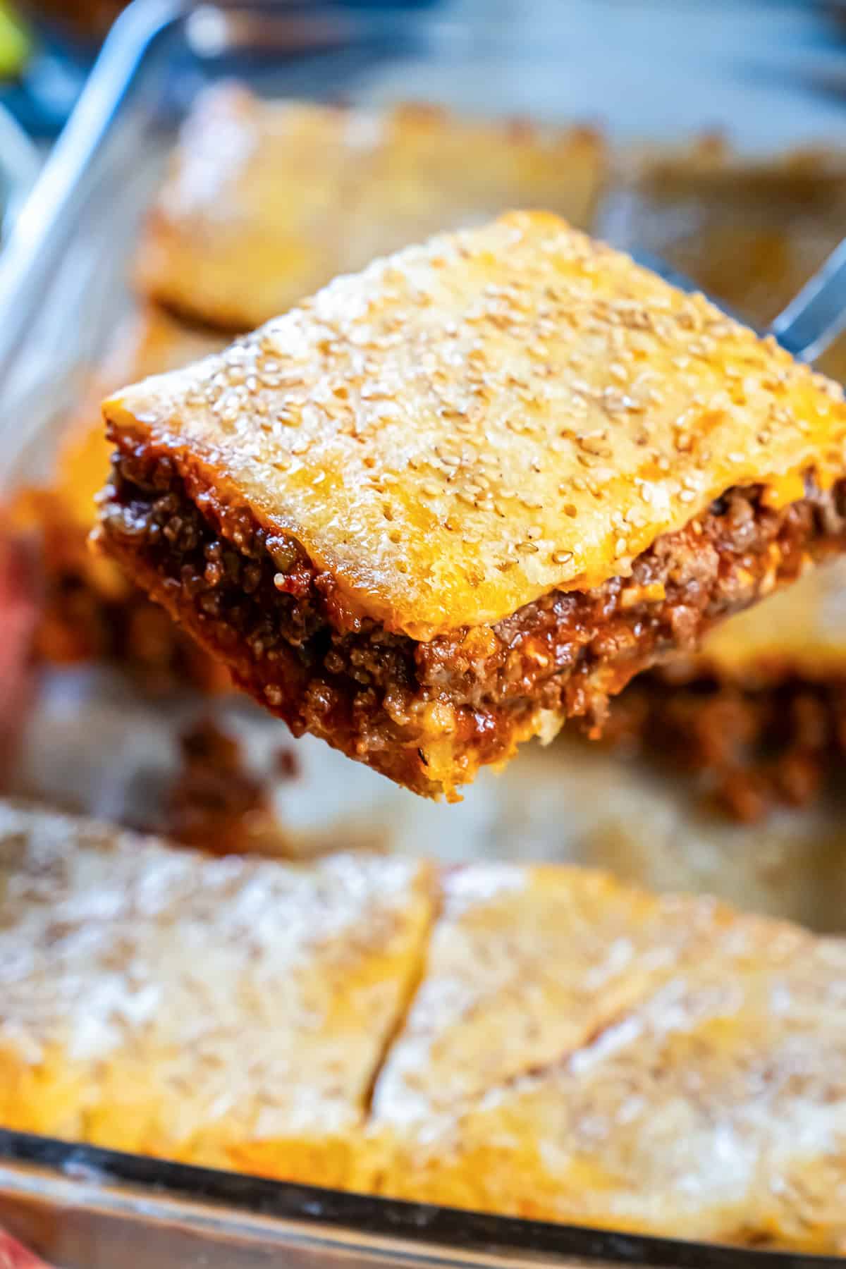 Sloppy Joe square being lifted from the pan, showing a golden, sesame-seed topped crescent roll crust and a thick, saucy beef filling.