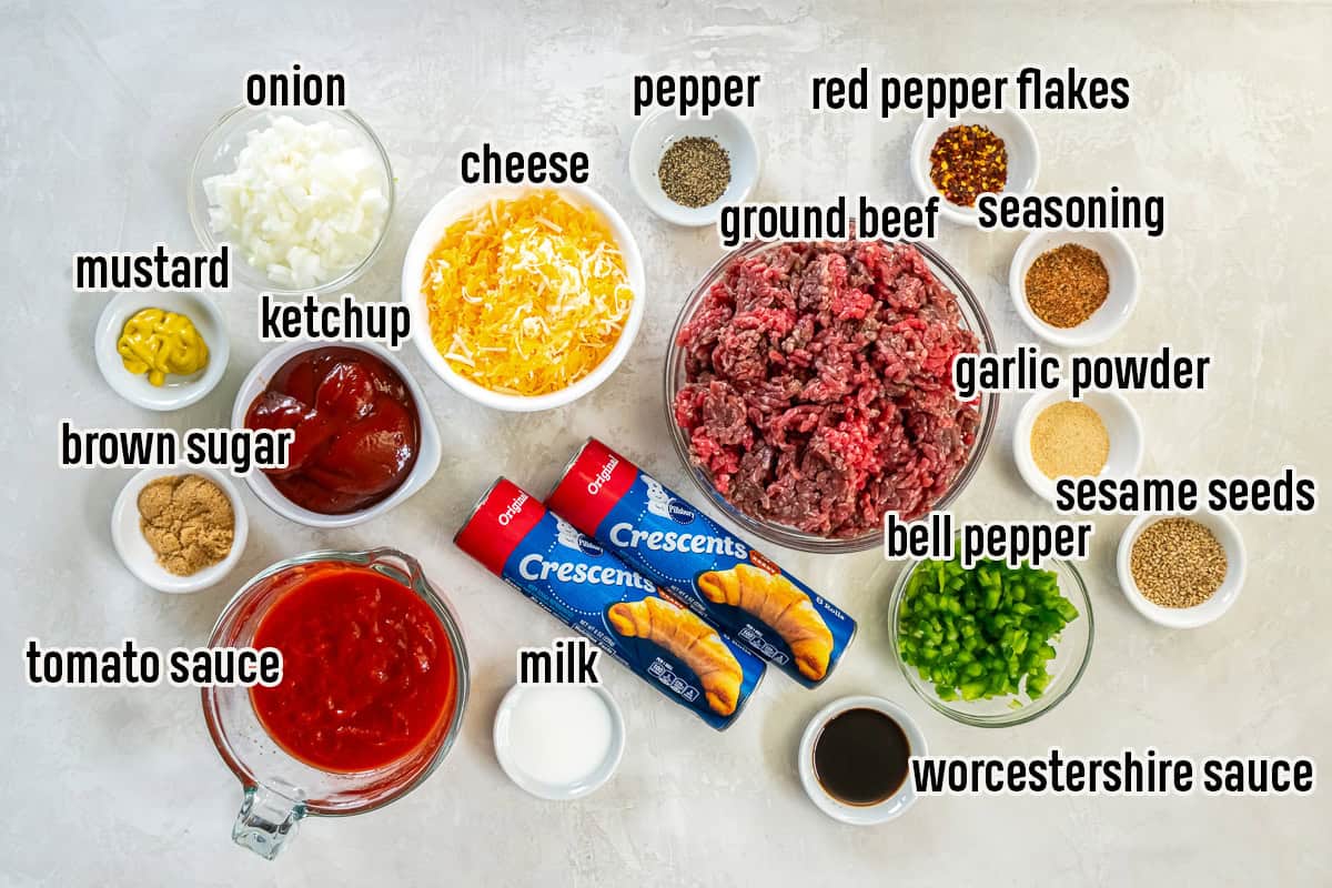Ingredients for sloppy joe squares including ground beef, diced onion, diced green bell pepper, shredded cheese, crescent roll dough, tomato sauce, ketchup, yellow mustard, Worcestershire sauce, brown sugar, Cajun seasoning, garlic powder, red pepper flakes, black pepper, milk, and sesame seeds arranged on a light surface.
