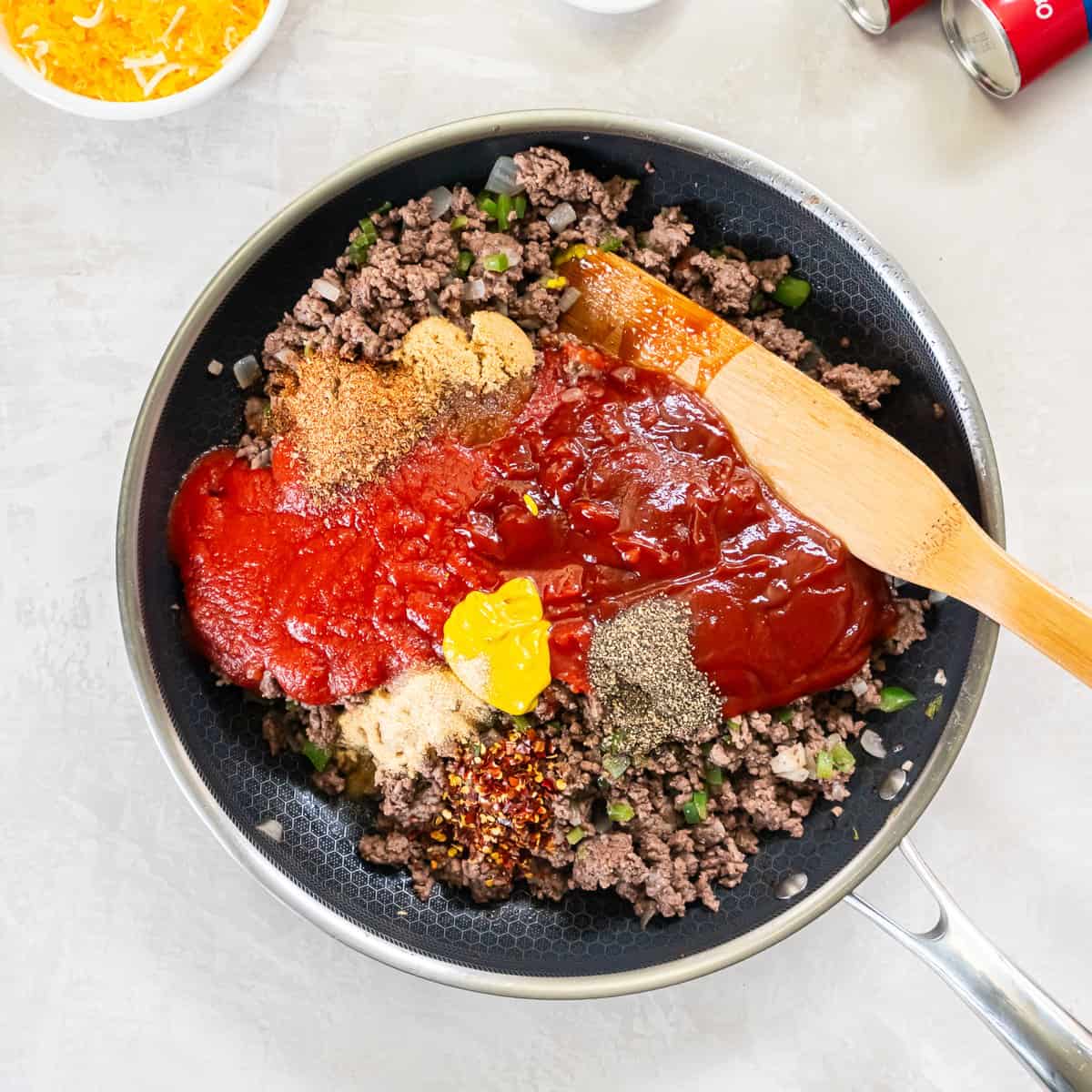 Tomato sauce, ketchup, mustard, brown sugar, and seasonings added to the cooked ground beef mixture in the skillet before stirring.