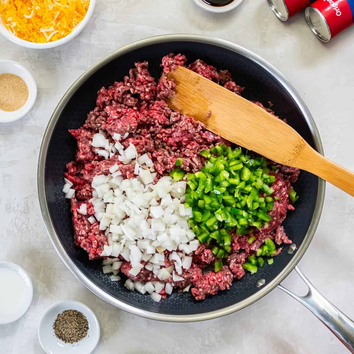 Ground beef in a large skillet with diced onion and green bell pepper just added, ready to be cooked and crumbled.