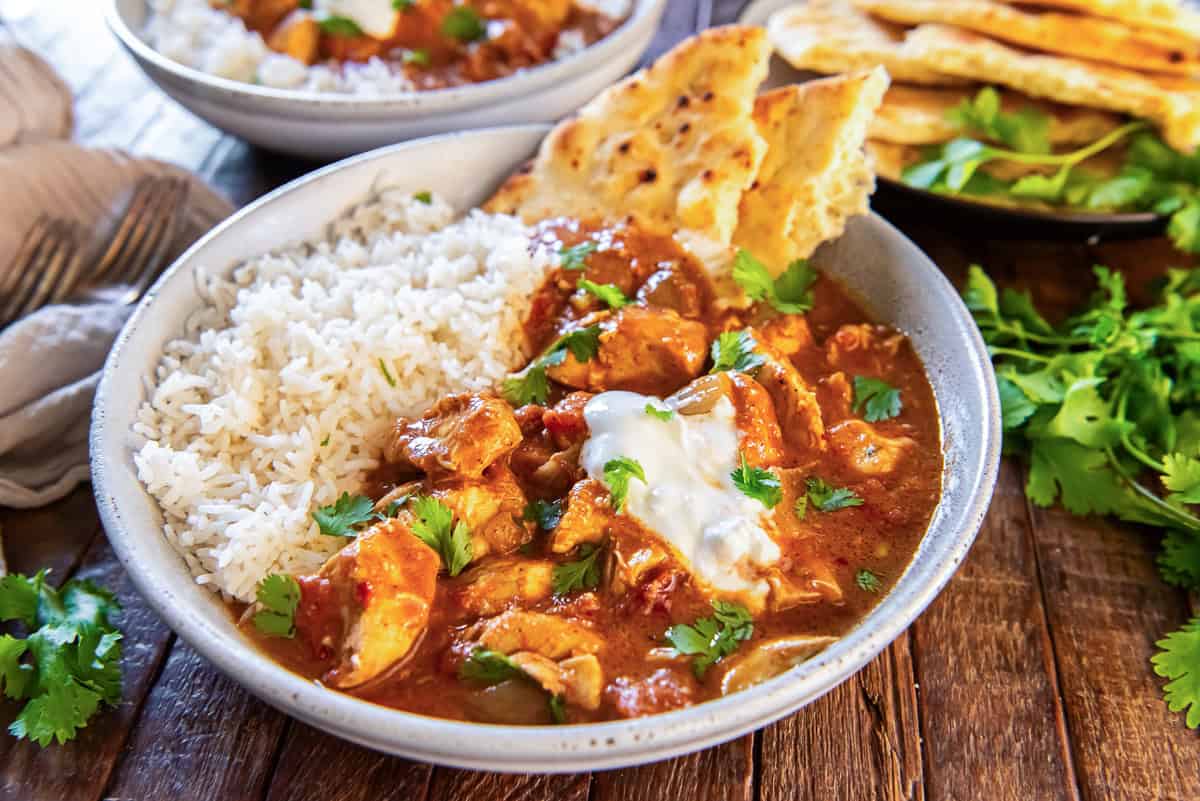 Bowl of slow cooker chicken tikka masala served with basmati rice and warm naan bread, garnished with fresh cilantro and a dollop of yogurt on a rustic wooden surface.