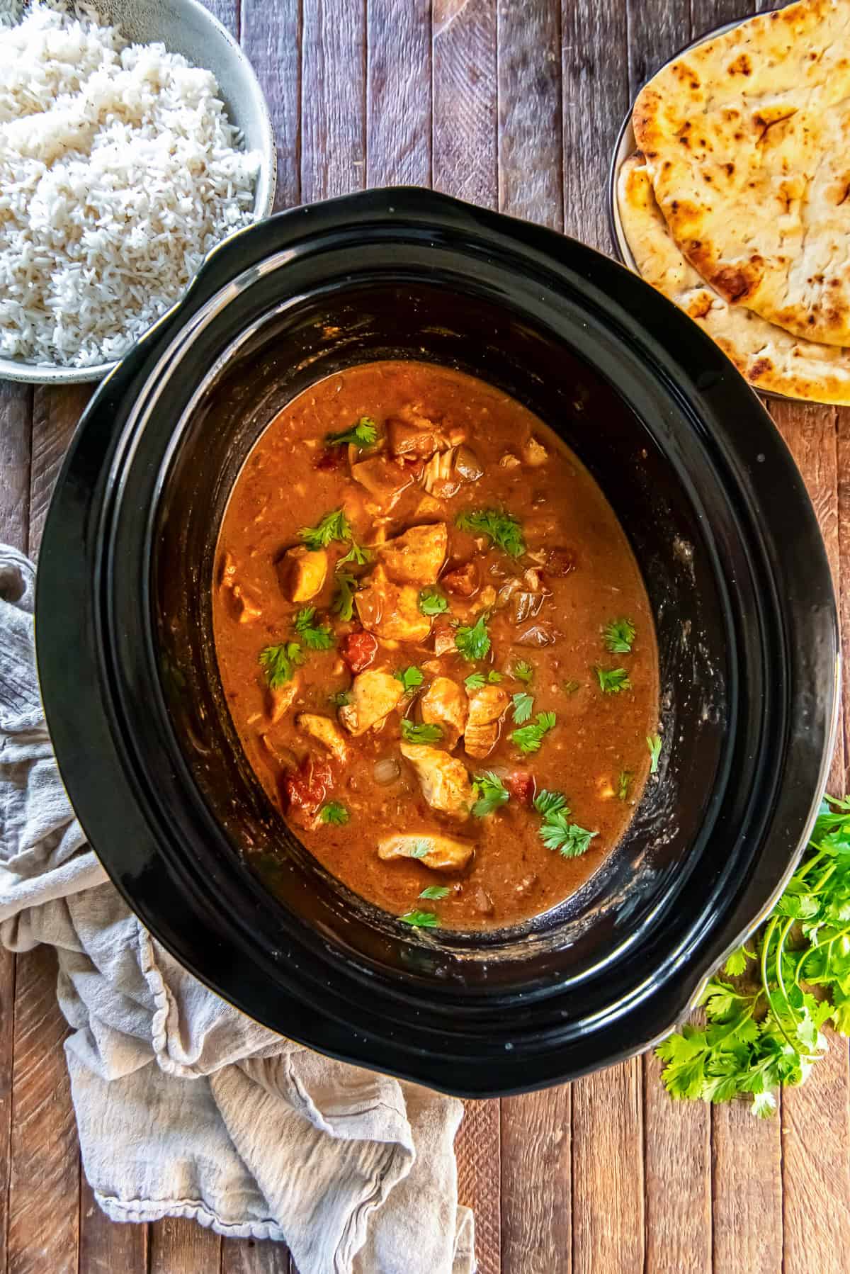 Chicken tikka masala in a slow cooker, garnished with cilantro and surrounded by bowls of basmati rice and warm naan bread.