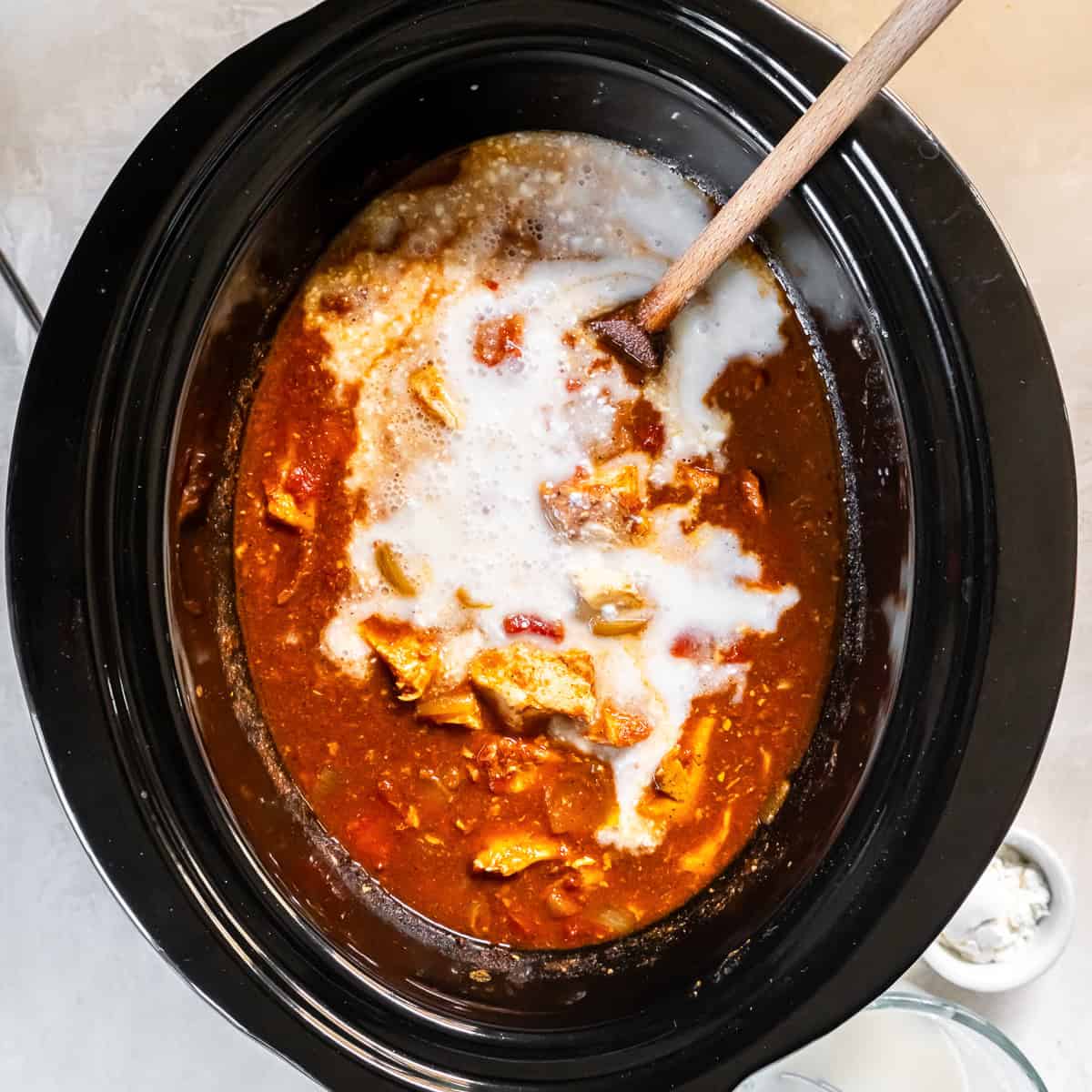Coconut milk being poured into the slow cooker over the chicken and tikka masala sauce before stirring.