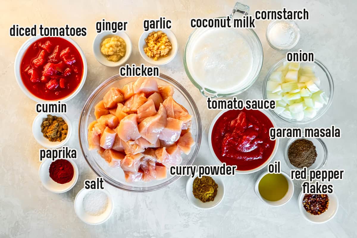 The ingredients for slow cooker chicken tikka masala arranged on a light surface, including cubed raw chicken, canned diced tomatoes, tomato paste, coconut milk, chopped onion, garlic, ginger, olive oil, curry powder, garam masala, paprika, red pepper flakes, salt, and cornstarch, with text labels identifying each ingredient.