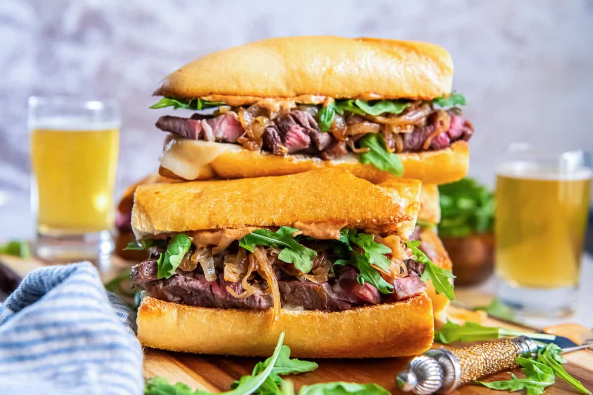 Stacked steak sandwiches on toasted baguette with sliced ribeye, melted cheese, caramelized onions, arugula, and smoky aioli on a wooden board with beer in the background.