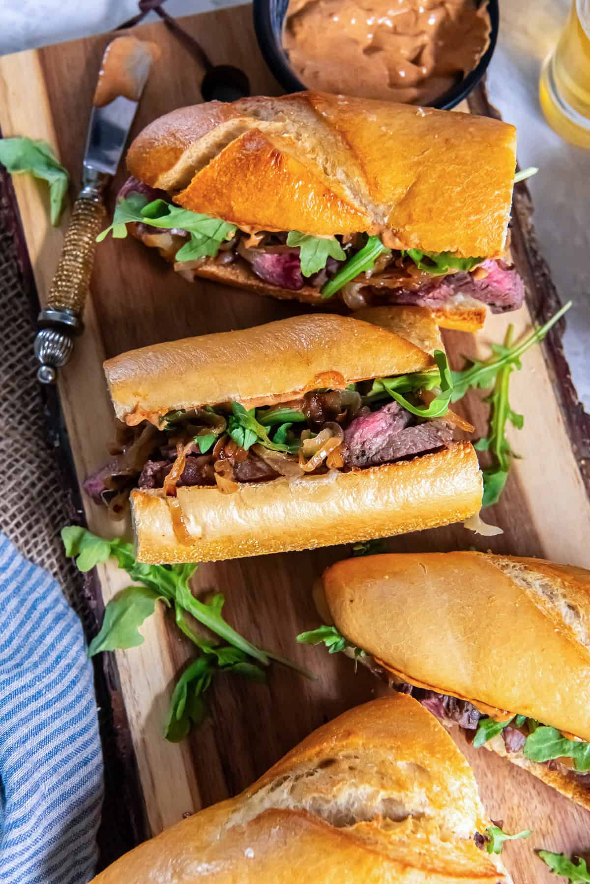 Steak sandwiches made with toasted baguette, sliced ribeye, caramelized onions, arugula, and smoky aioli arranged on a wooden cutting board.