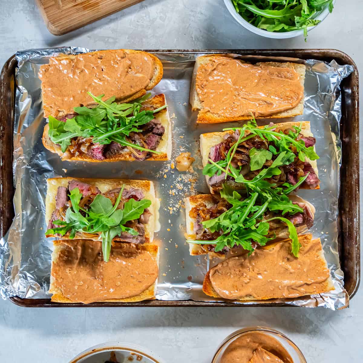 Smoky aioli spread on toasted baguette halves with sliced steak, caramelized onions, and fresh arugula added on top, arranged on a foil-lined baking sheet.