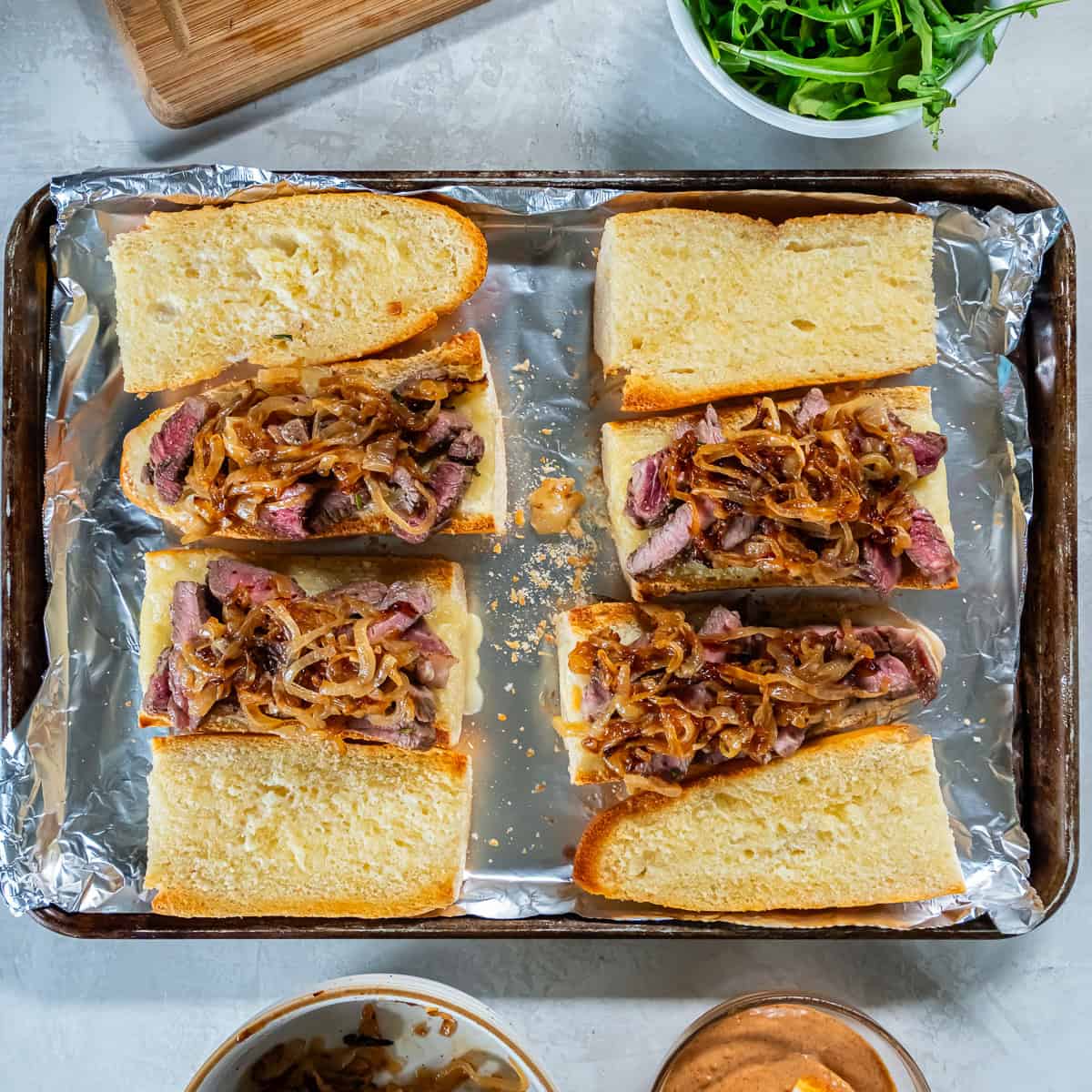 Toasted baguette pieces topped with sliced ribeye and caramelized onions, ready to be finished with sauce and greens.