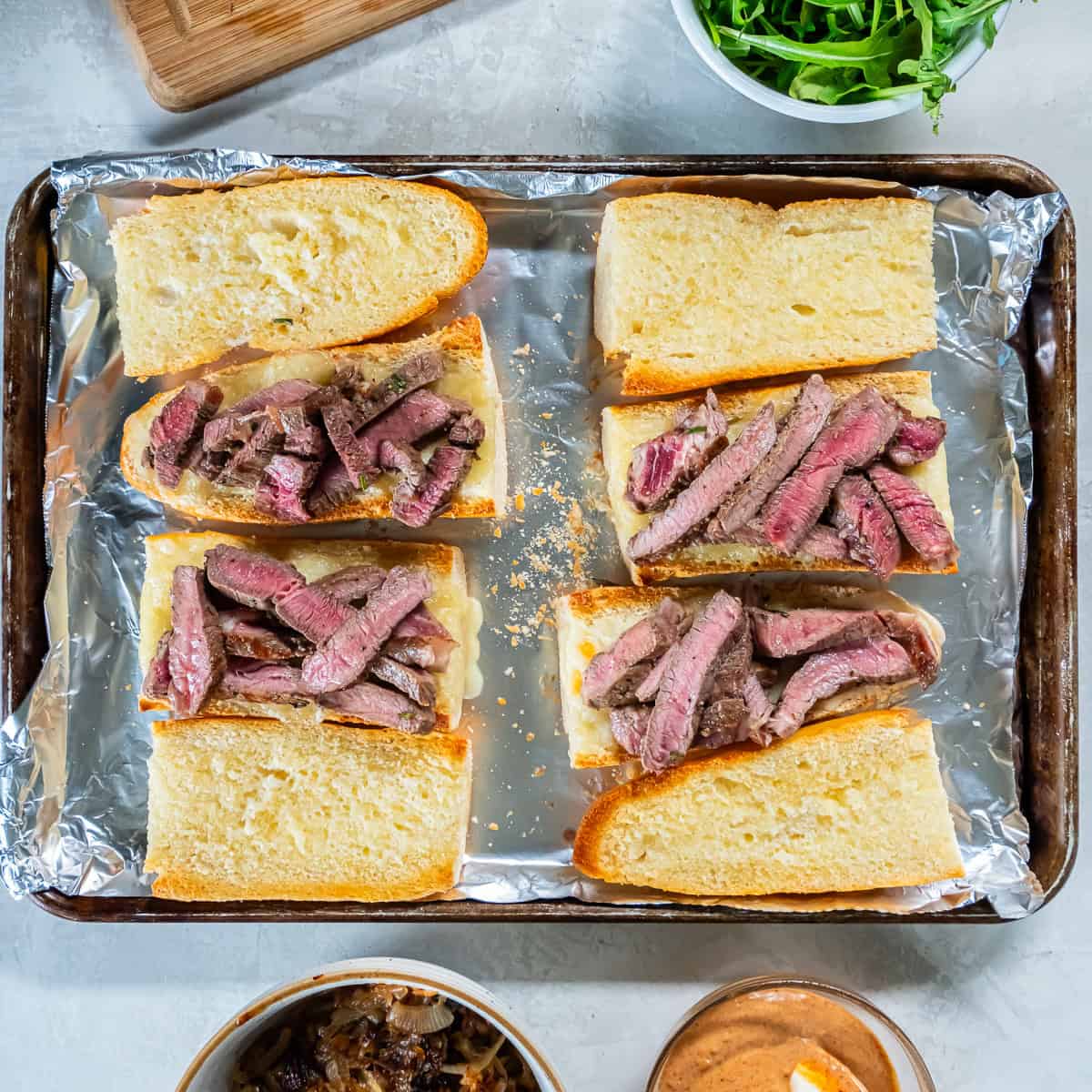 Sliced medium-rare ribeye layered onto toasted baguette pieces on a foil-lined baking sheet.