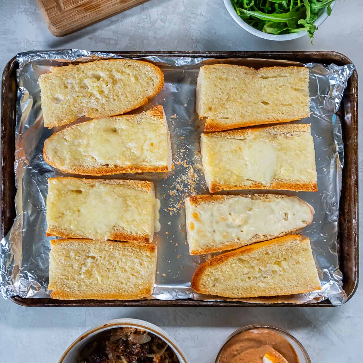 Toasted baguette halves topped with melted Havarti cheese, arranged on a foil-lined baking sheet and ready for the steak and toppings.