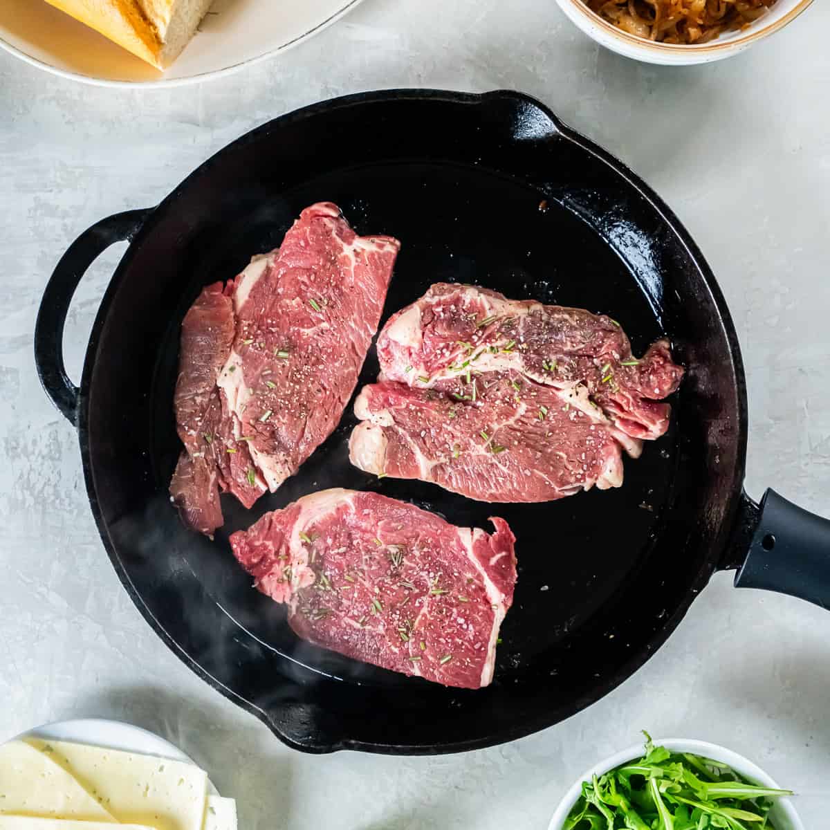 Thin-sliced boneless ribeye steaks seasoned with salt, pepper, and rosemary cooking in a cast iron skillet.