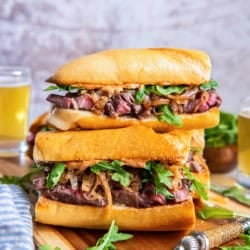 Stacked steak sandwiches with toasted baguette, sliced ribeye, melted cheese, caramelized onions, arugula, and smoky aioli on a wooden cutting board with glasses of beer in the background.
