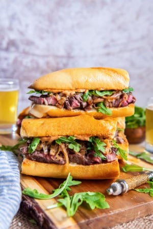 Stacked steak sandwiches with toasted baguette, sliced ribeye, melted cheese, caramelized onions, arugula, and smoky aioli on a wooden cutting board with glasses of beer in the background.