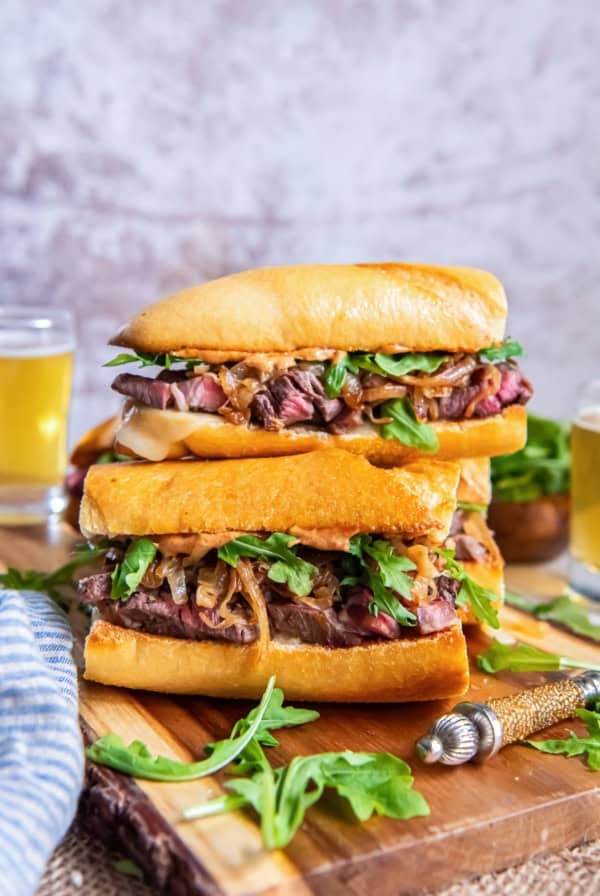 Stacked steak sandwiches with toasted baguette, sliced ribeye, melted cheese, caramelized onions, arugula, and smoky aioli on a wooden cutting board with glasses of beer in the background.