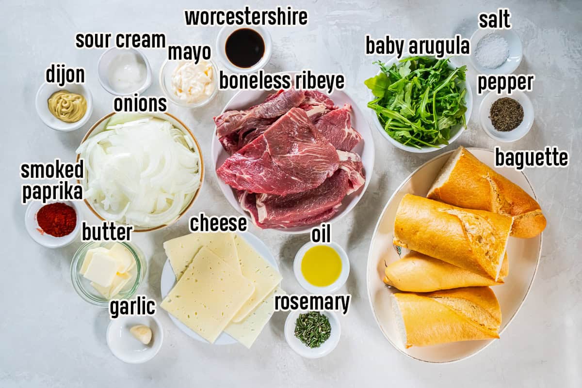 Ingredients for a steak sandwich including thin sliced boneless ribeye, baguette pieces, Havarti cheese, sliced onions, baby arugula, mayonnaise, sour cream, Dijon mustard, Worcestershire sauce, butter, garlic, olive oil, fresh rosemary, smoked paprika, salt, and black pepper.
