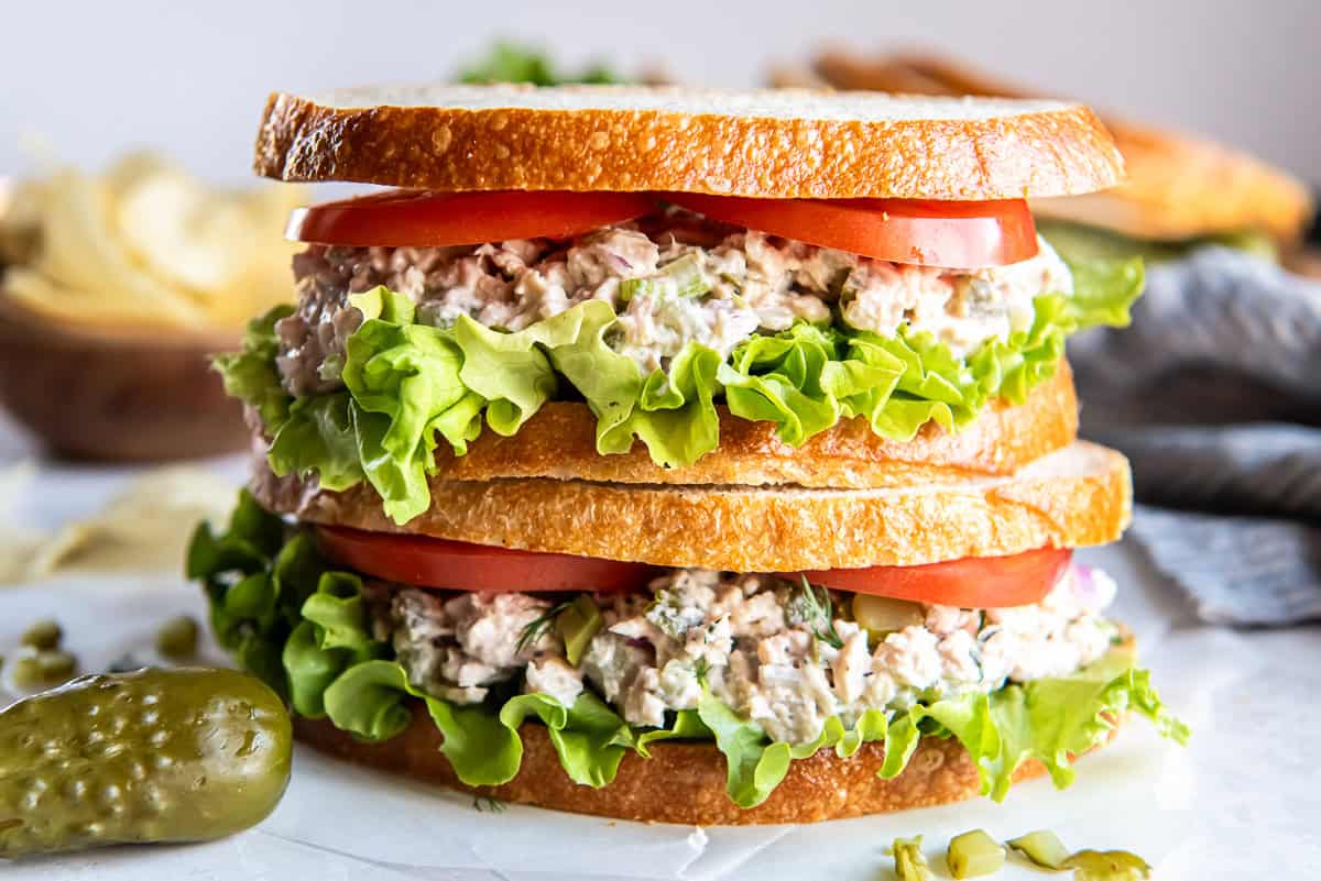 Dill pickle tuna salad sandwich layered with lettuce and tomato on sliced bread, served with potato chips and a pickle on the side.